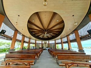 Interior view of an open-air circular chapel featuring wooden pews, a central altar with a crucifix, and a large dome ceiling with radial beams.