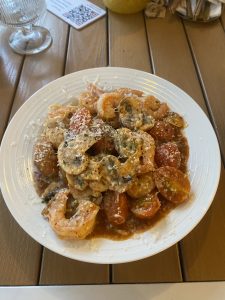A white plate of shrimp with mushrooms and cherry tomatoes in a savory sauce, topped with grated cheese and herbs, served on a wooden outdoor table beside a glass and a QR code menu card.