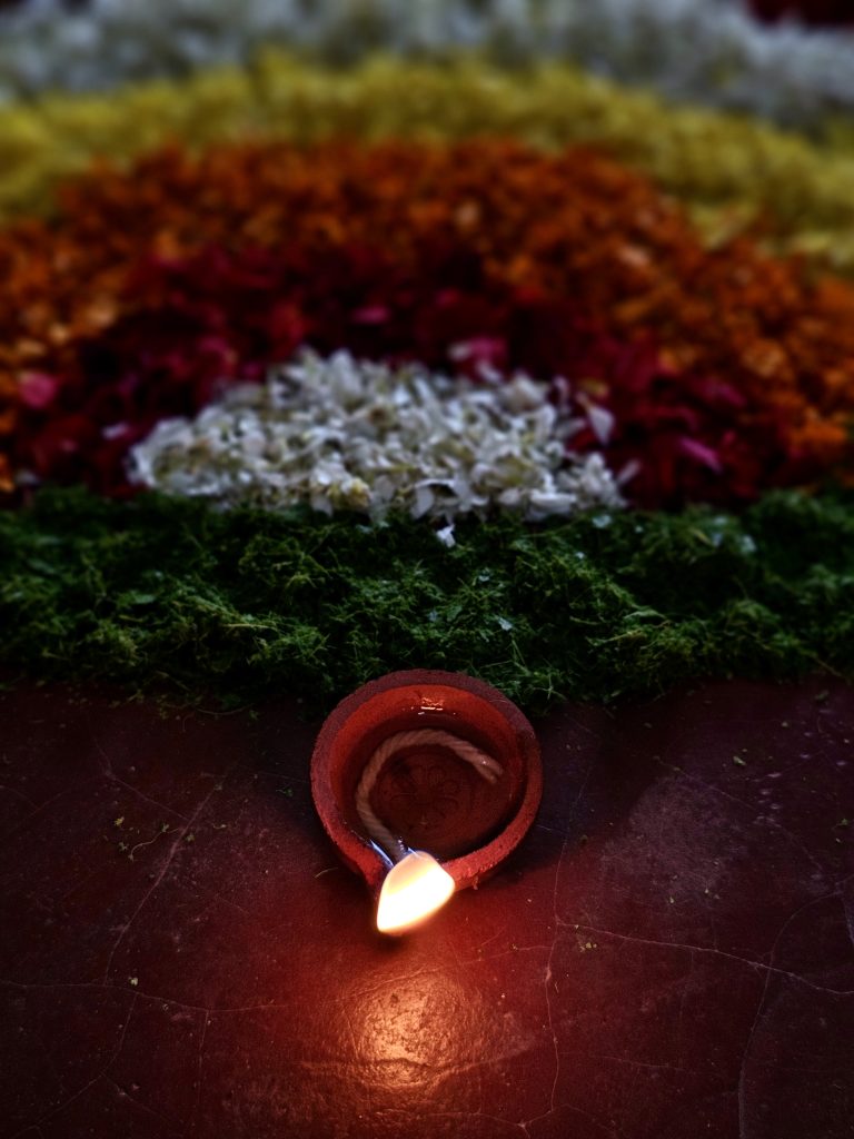 A small traditional clay oil lamp glows warmly in front of colorful flower decorations. The soft focus and dark background highlight the lamp and its gentle flame, creating a peaceful festive scene. Captured in Perumanna, Kozhikode, Kerala.