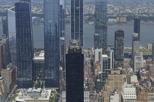 View of modern glass skyscrapers with the Hudson River in the background. Tall office towers line a busy avenue, while the river and distant cityscape stretch across the horizon under a hazy sky