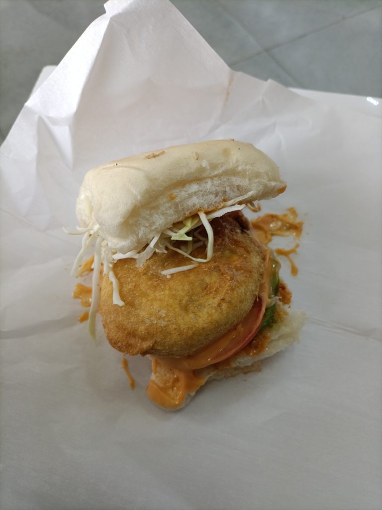 Italian-style vada pav with a fried patty, tomato, cabbage, and creamy sauce in a bun.