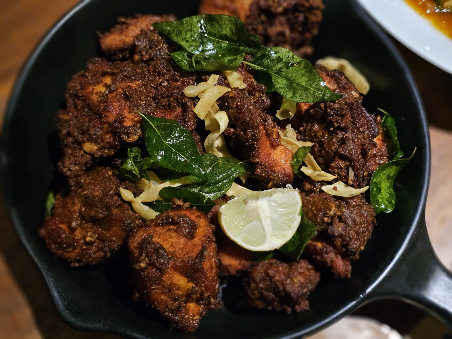 A close-up photograph of crispy Kerala-style fried chicken served in a black dish. Fresh curry leaves and lime slices garnish the spicy-looking chicken, while the dark background and shallow focus give the dish a rich, appetizing look. Location: Perumanna, Kozhikode, Kerala.