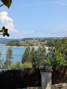 Scenic coastal landscape of a Galician estuary bay with lush green trees, a sandy beach, and village houses scattered along the distant hillside under a summer sky.