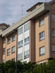 Modern residential apartment building with beige facade, enclosed glass balconies, and rooftop antennas against a cloudy sky.