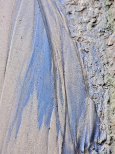 A close-up view of sand left behind after a flood shows flowing blue and grey patterns, fine textures, and patches of wet, compacted sand. The floodwater has created an unusual natural abstract pattern of lines and shapes. Captured in Koolimadu, Kozhikode, Kerala.