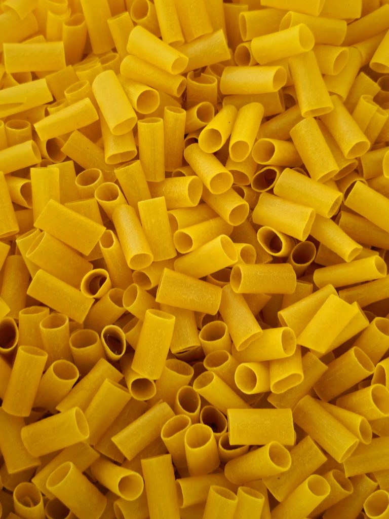 A close-up view shows many short, hollow pieces of yellow tube-shaped pasta (Macaroni) arranged in a dense pile. The repeated shapes, warm color, and simple composition create an interesting food texture. Captured in Poovattuparamba, Kozhikode, Kerala.