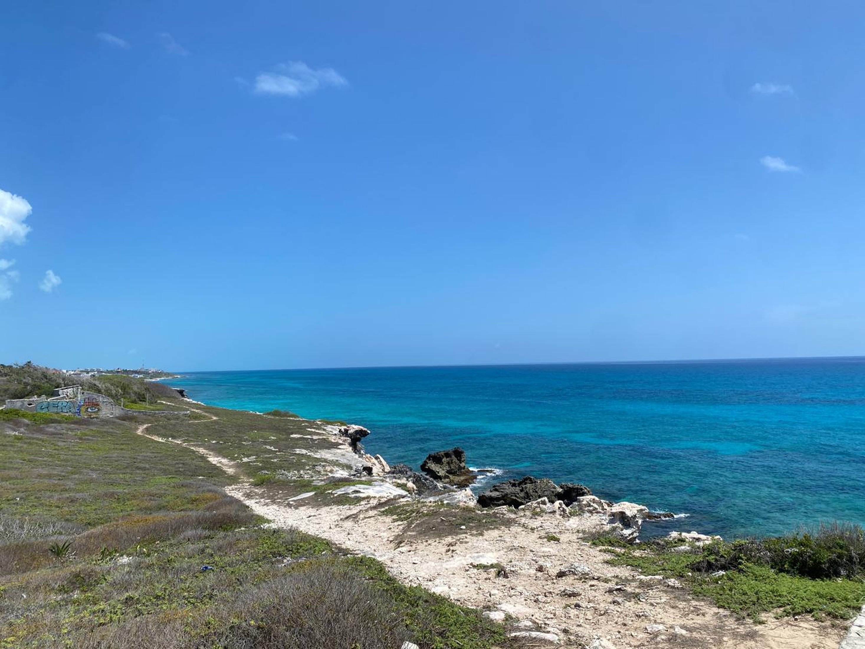 Panoramic view of a rugged coastal landscape with rocky cliffs, low vegetation, turquoise ocean water, and a bright blue sky with scattered white clouds.