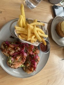 A plate of open-faced toast topped with salmon, avocado spread, pickled red onions, sesame seeds, and scallions, served with a basket of french fries and a small dish of ketchup on a wooden table.