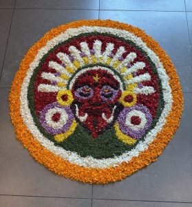 A colorful Pookkalam floral arrangement with a red face design, surrounded by vibrant petals on a tiled floor.