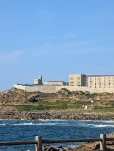 Coastal stone fortress and Parador de Baiona hotel sitting atop a rocky hill overlooking blue ocean waves in Galicia, Spain.