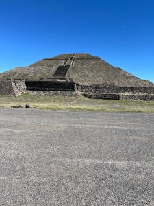 A pyramid build in the Aztec style.  Seen from about 100 meters away. The sky above is clear blue and the moon appears very small.  In the foreground is a wide road.