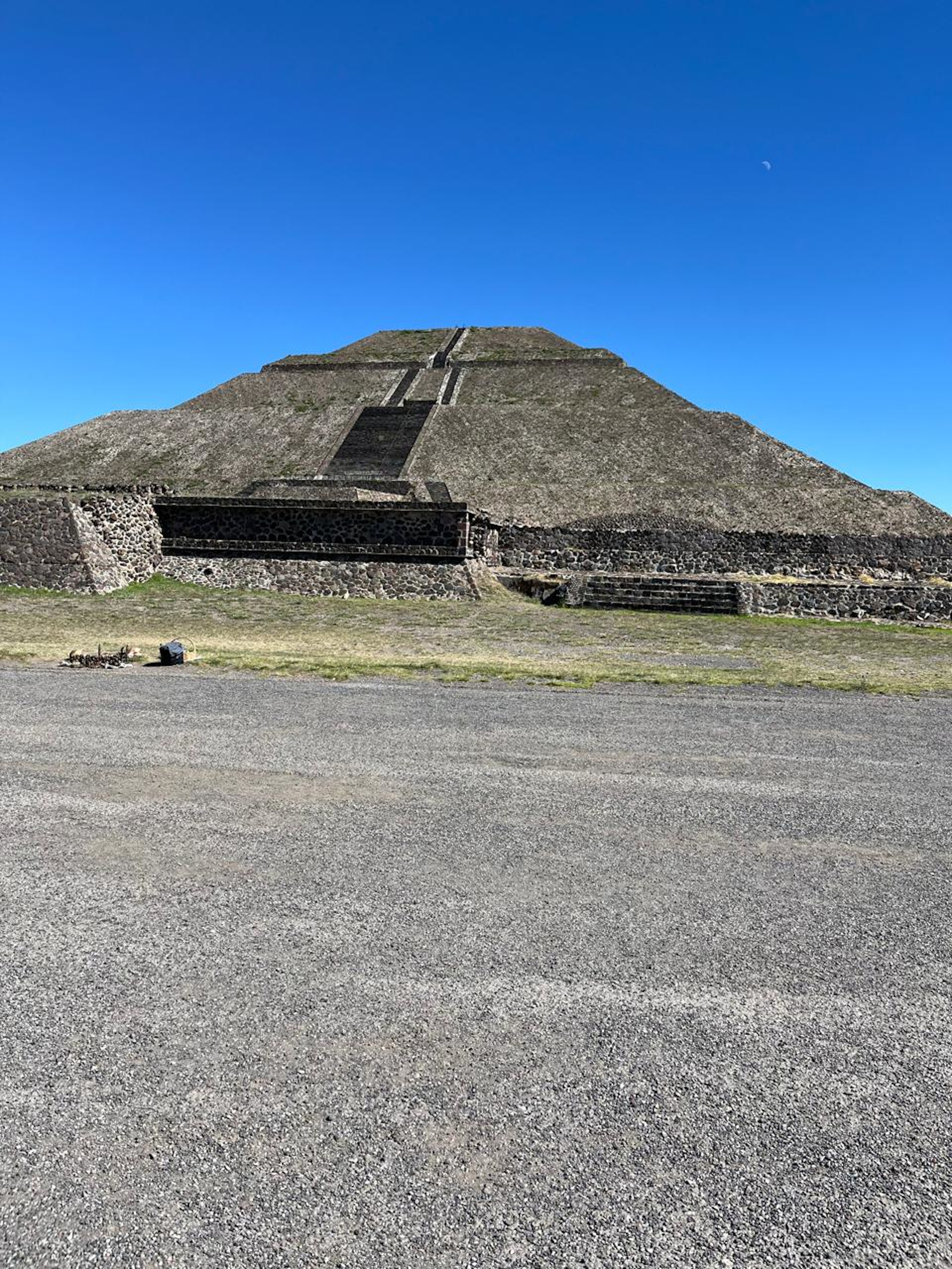 A pyramid build in the Aztec style. Seen from about 100 meters away. The sky above is clear blue and the moon appears very small. In the foreground is a wide road.