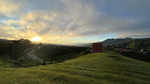 Sunset over a lush alpine village, with a red barn, winding road, distant mountains, and dramatic clouds creating a peaceful rural scene.