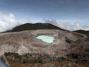 A panoramic view of a volcanic crater featuring a light green crater lake at the center, surrounded by rocky, gray terrain with various textures and colors, including reddish and yellowish hues.