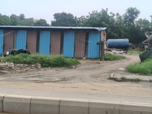 A dirt path through an overgrown area with small wooden structures, a large blue water tank, debris, and patches of grass.