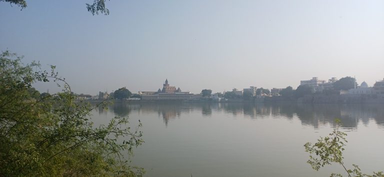 A calm lake reflecting buildings and trees on a peaceful morning under a clear sky.