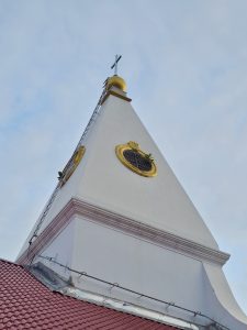 A close-up upward view of a white church tower with a golden dome and cross at the top. The simple architecture stands clearly against the pale blue sky, while the tiled roof and decorative details add interest to the lower part of the photograph. Location: Mahé, Puducherry.