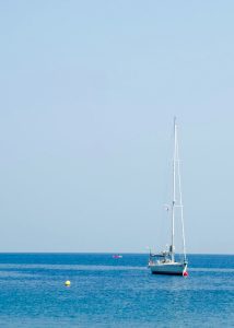 White sailboat anchored on calm blue water, with a yellow buoy in the foreground and a small boat in the distance.
