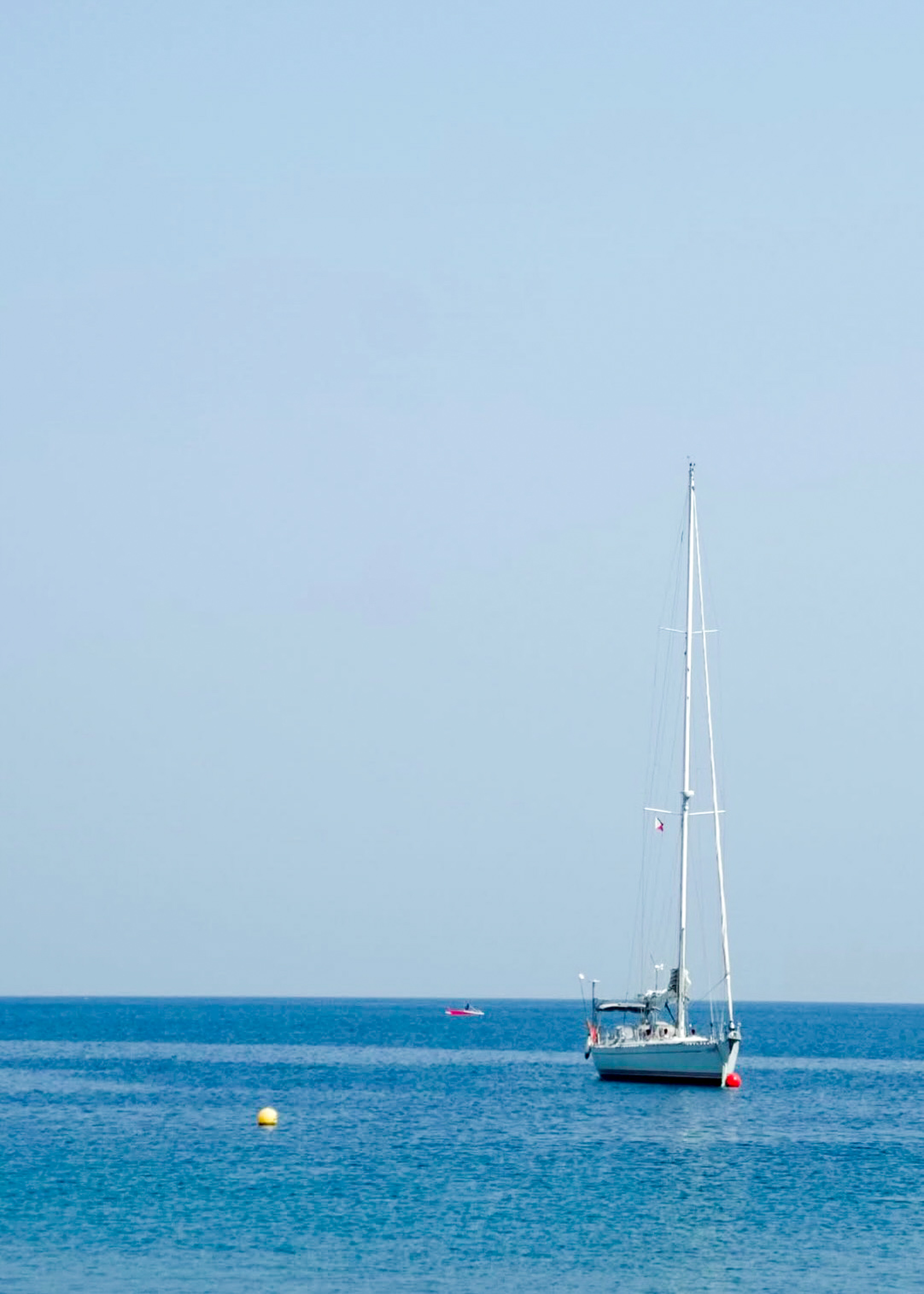 White sailboat anchored on calm blue water, with a yellow buoy in the foreground and a small boat in the distance.