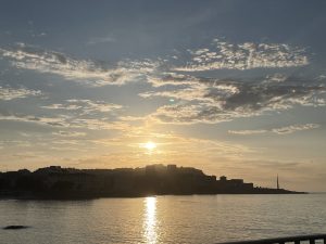 
Sunset over a calm coast with silhouetted hills, clouds, and reflections on the water.