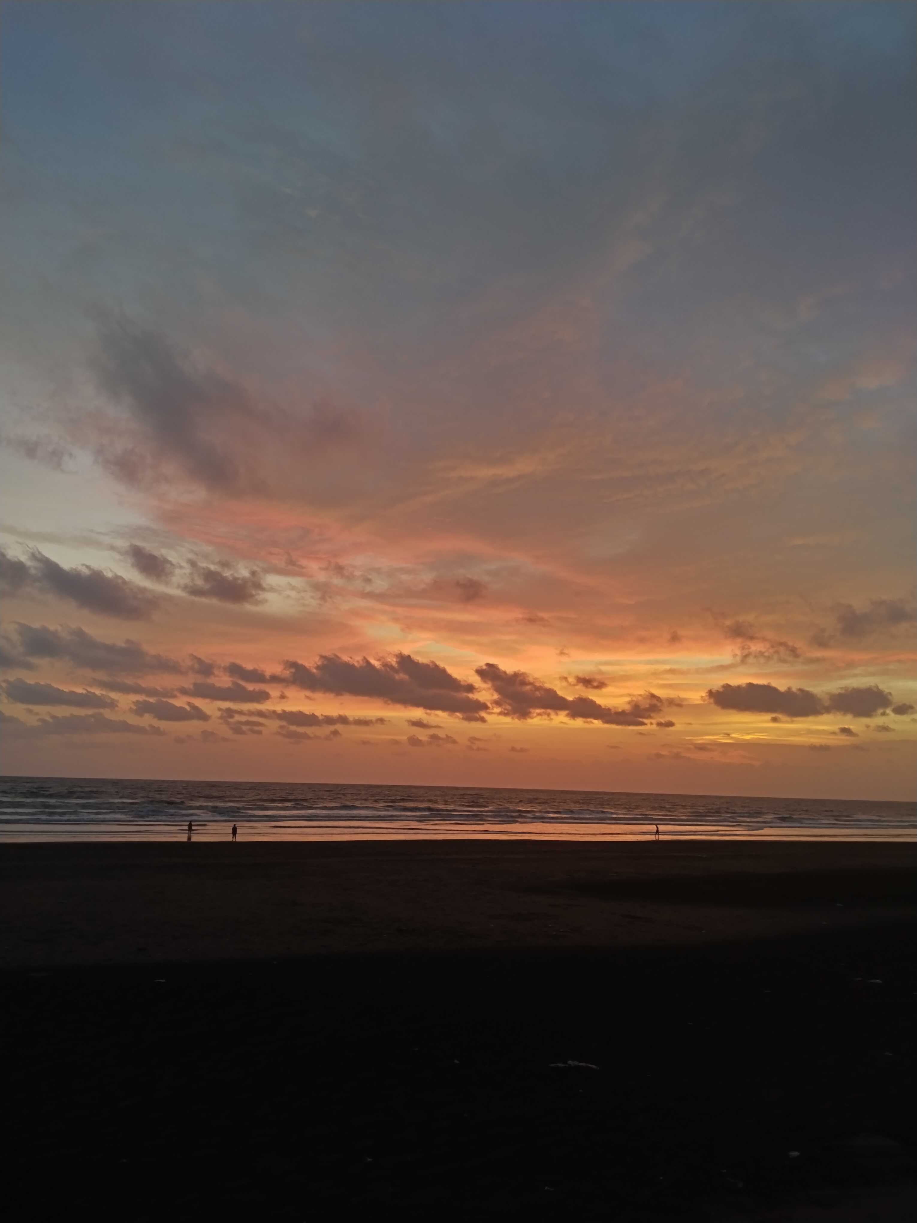 A colorful sunset over the ocean with orange, pink, and purple clouds reflecting across the water, as a few people walk along the dark sandy beach in silhouette.