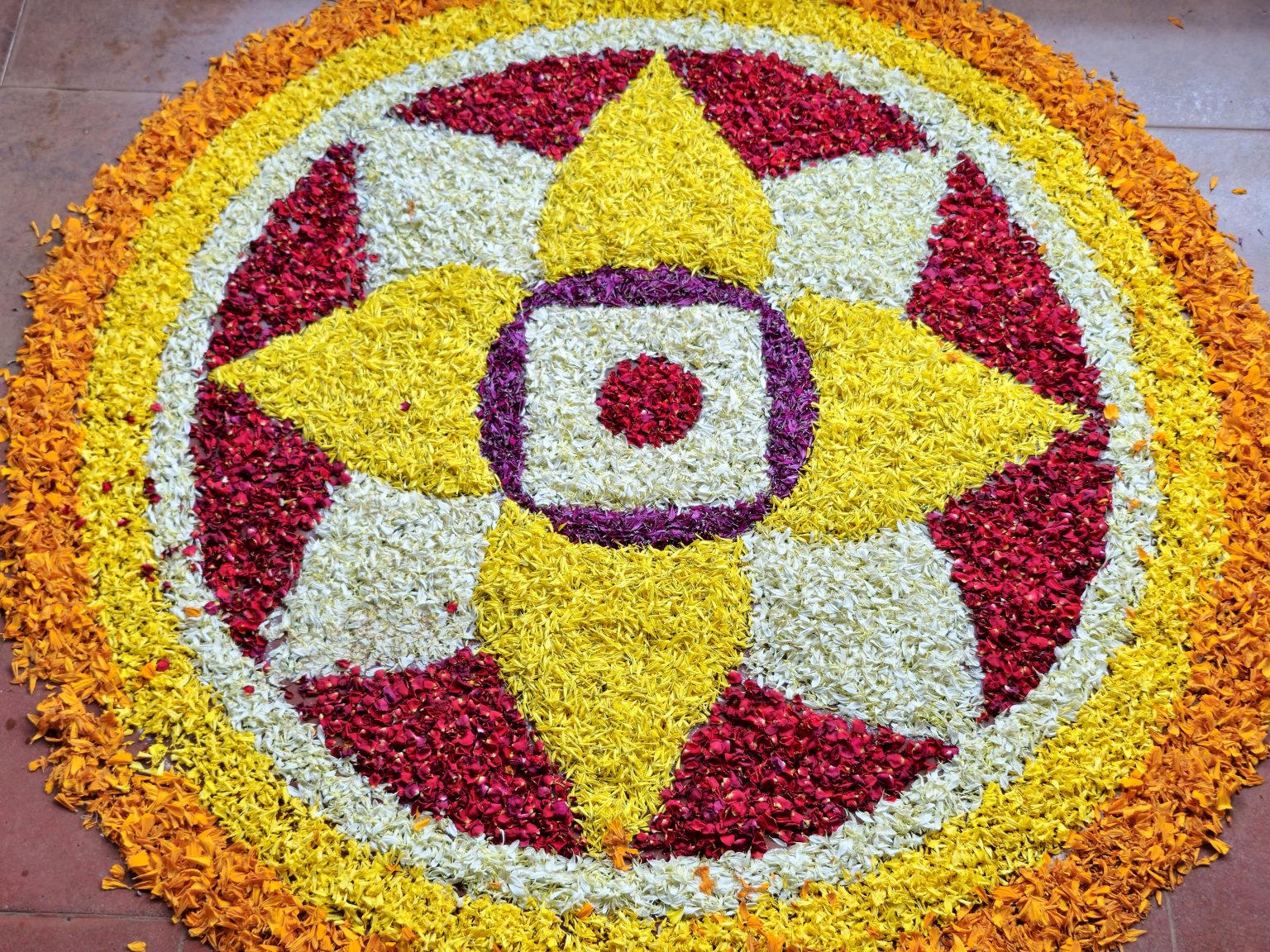 A colorful circular Pookkalam made with fresh flower petals, featuring bright yellow, red, white, and orange patterns arranged in a detailed geometric design. The close-up view clearly shows the texture and colors of the flowers. Location: Perumanna, Kozhikode, Kerala.