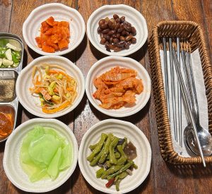 Assorted Korean side dishes in small white bowls on a wooden table, with kimchi, beans, vegetables, peppers, and utensils.