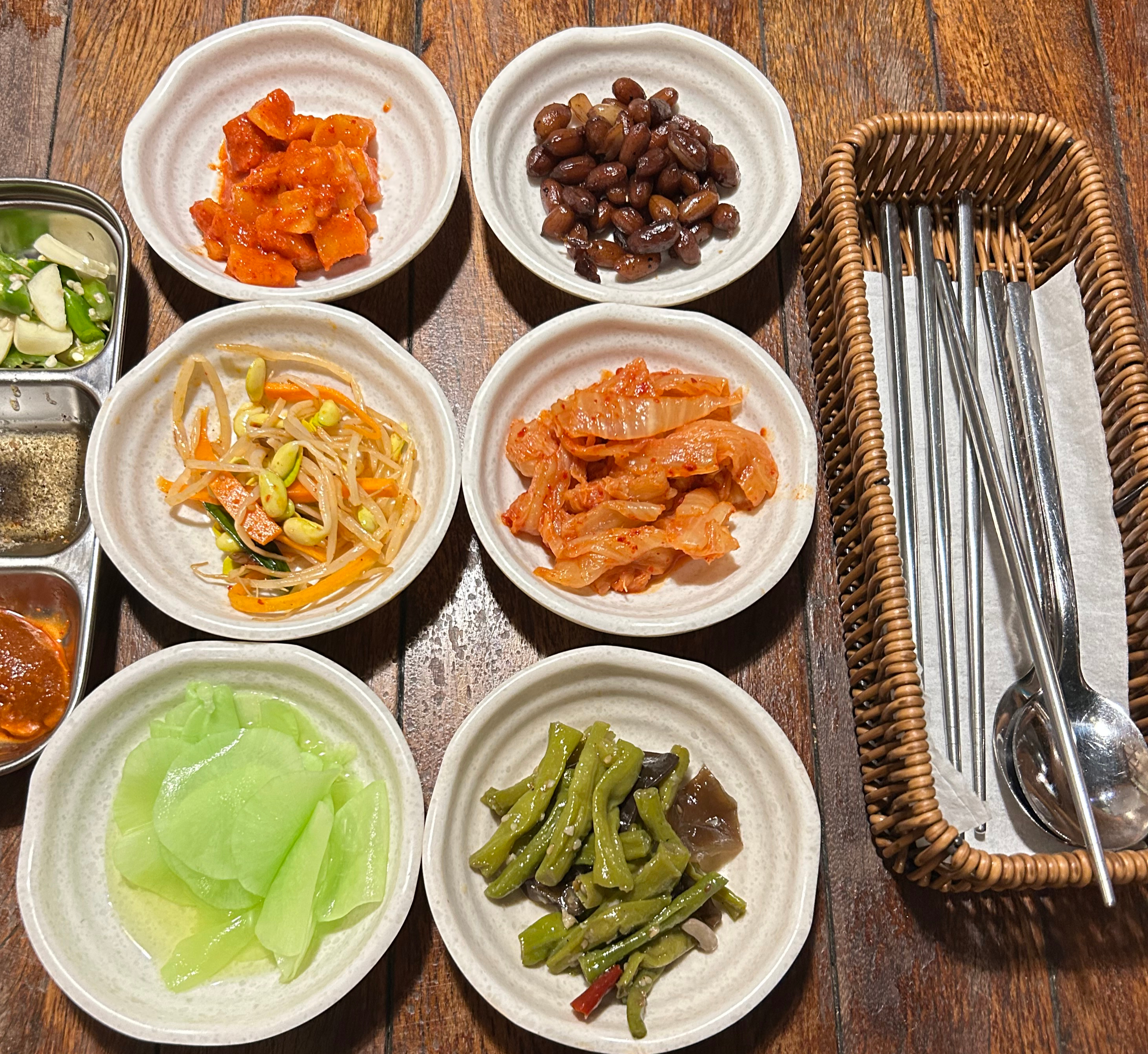 Assorted Korean side dishes in small white bowls on a wooden table, with kimchi, beans, vegetables, peppers, and utensils.