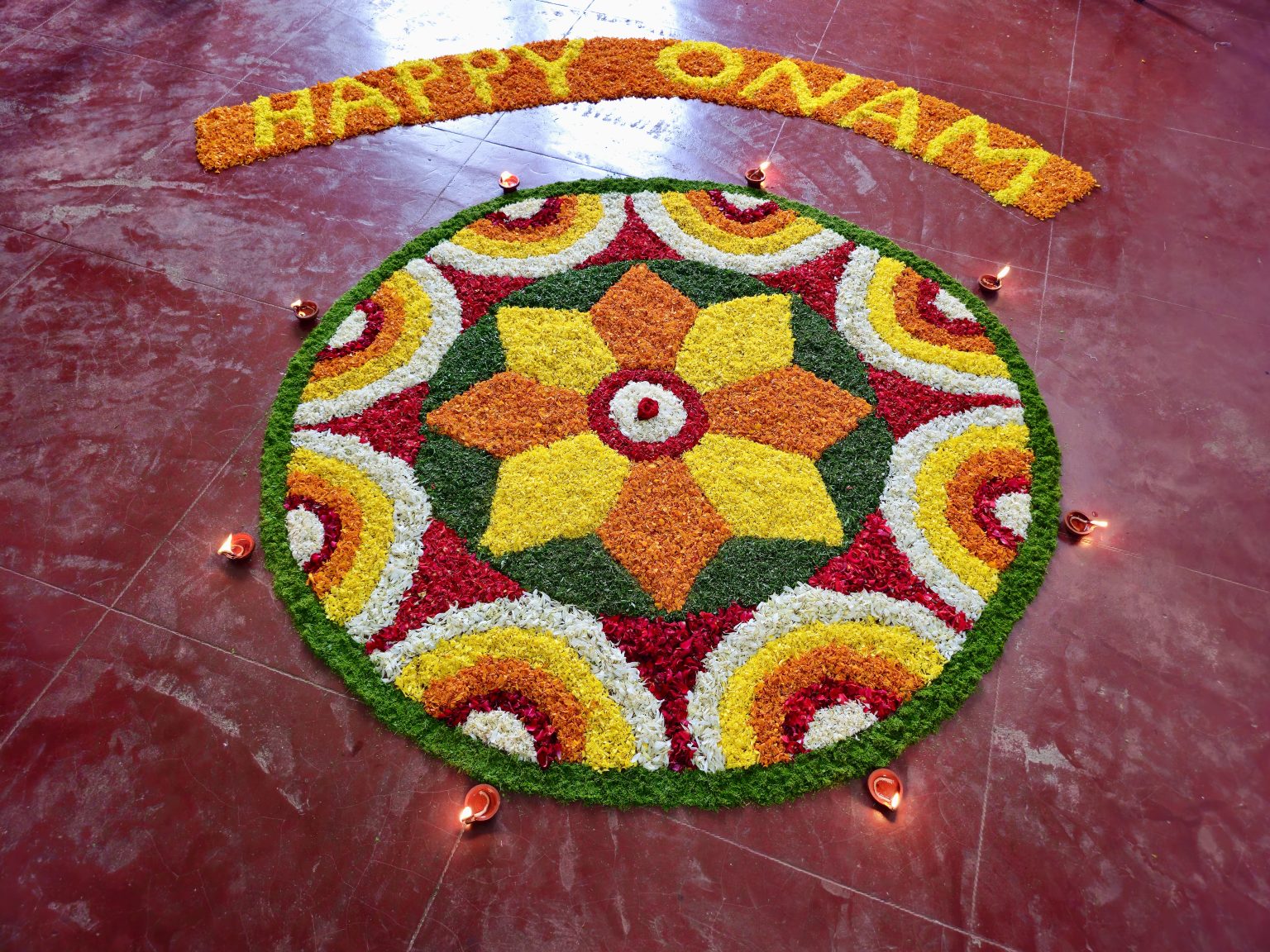 A vibrant circular Pookkalam featuring layers of red, yellow, orange, white, and green flower petals. Traditional oil lamps surround the design, adding warmth and a festive feel. The symmetrical composition makes the colorful floral pattern stand out. Location: Perumanna, Kozhikode, Kerala.
