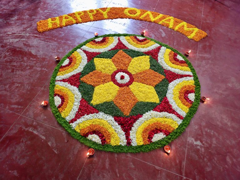 A vibrant circular Pookkalam featuring layers of red, yellow, orange, white, and green flower petals. Traditional oil lamps surround the design, adding warmth and a festive feel. The symmetrical composition makes the colorful floral pattern stand out. Location: Perumanna, Kozhikode, Kerala.