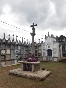 Galician-style cemetery with a stone cross decorated with bright pink flowers and rows of burial niches under a cloudy sky.