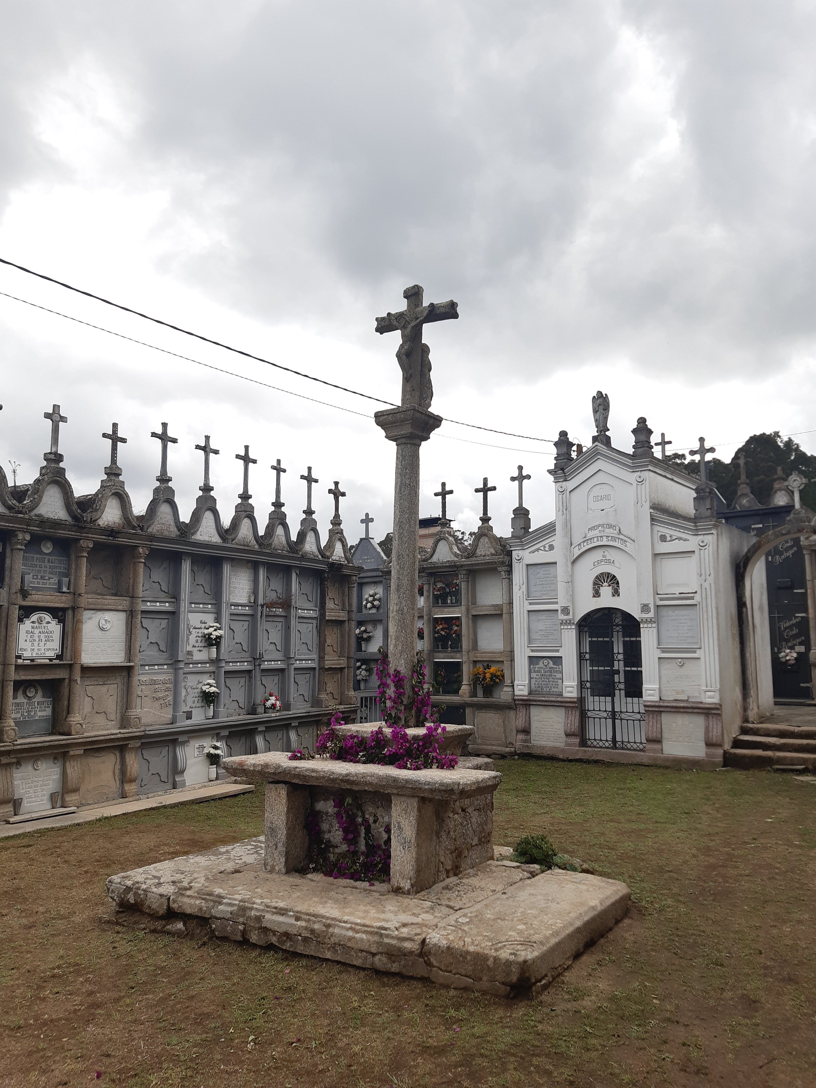 Galician-style cemetery with a stone cross decorated with bright pink flowers and rows of burial niches under a cloudy sky.