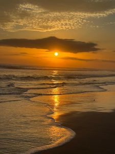 Sunset over the ocean with the sun behind dark clouds, reflecting on gentle waves.