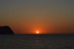 The sun sets on the horizon over a calm ocean, casting warm orange and gold light across the water.