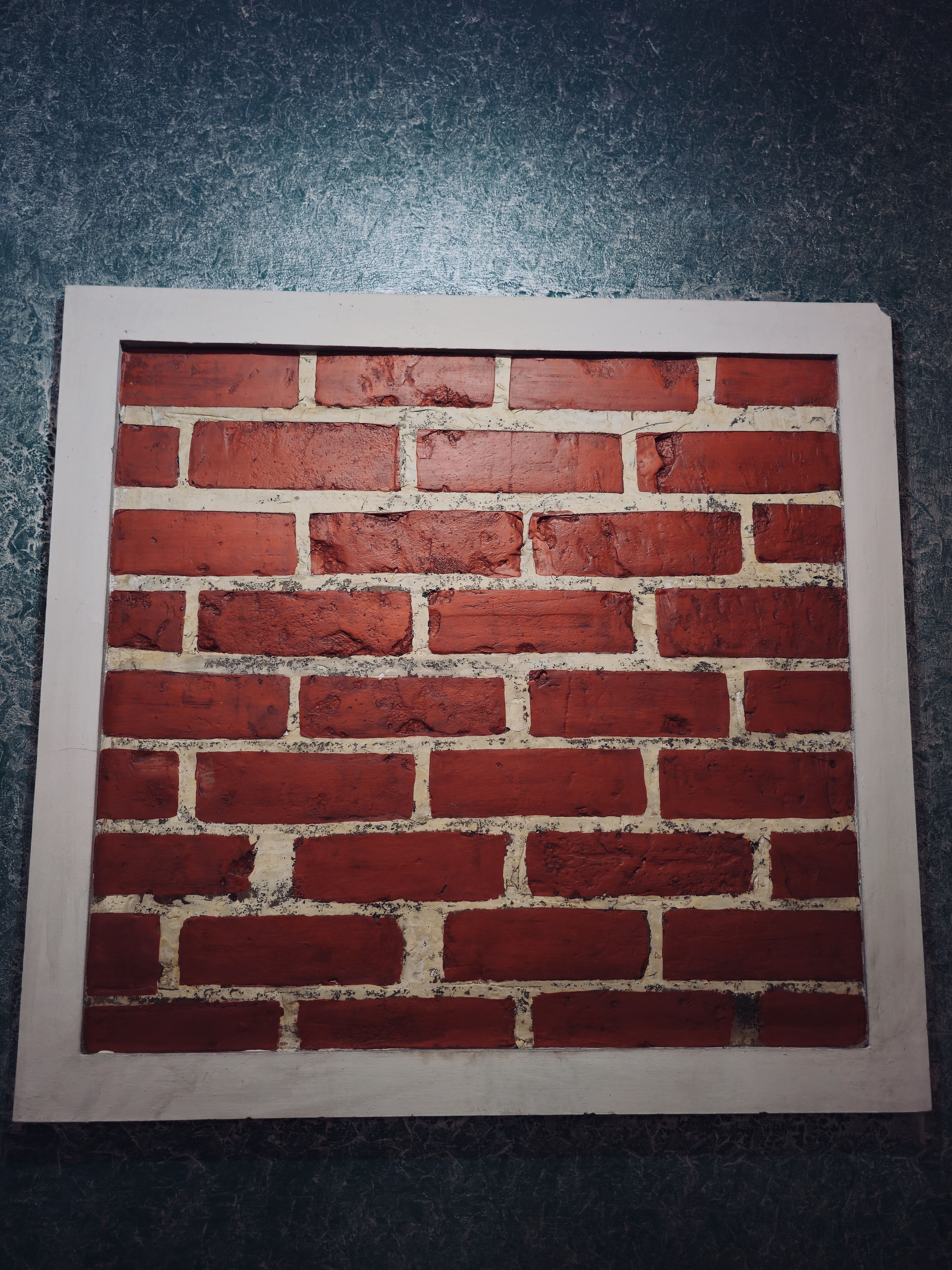 A rectangular panel displays rows of red bricks with uneven textures and pale mortar lines, framed by a dark blue textured wall. The simple geometric pattern creates an interesting study of color, texture, and repetition. Captured in Chalappuram, Kozhikode, Kerala.