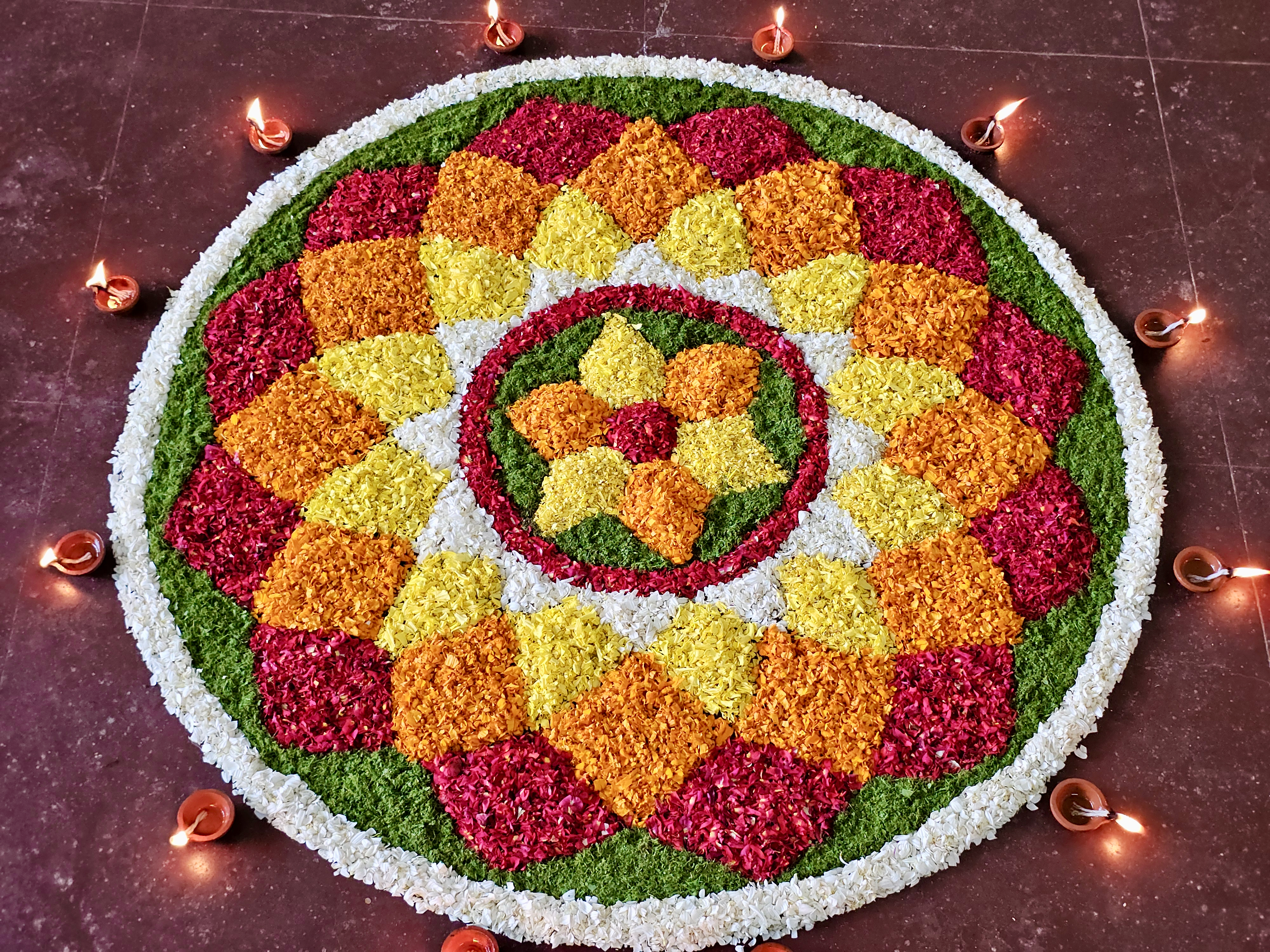 A beautifully arranged circular Pookkalam made from colorful flower petals, surrounded by small traditional oil lamps. The flower patterns and warm lamps create a festive atmosphere associated with Onam celebrations. Location: Perumanna, Kozhikode, Kerala.