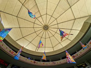 Low-angle shot looking directly up at a large fabric dome canopy ceiling with colorful triangular banners suspended beneath it inside a circular shopping mall interior.