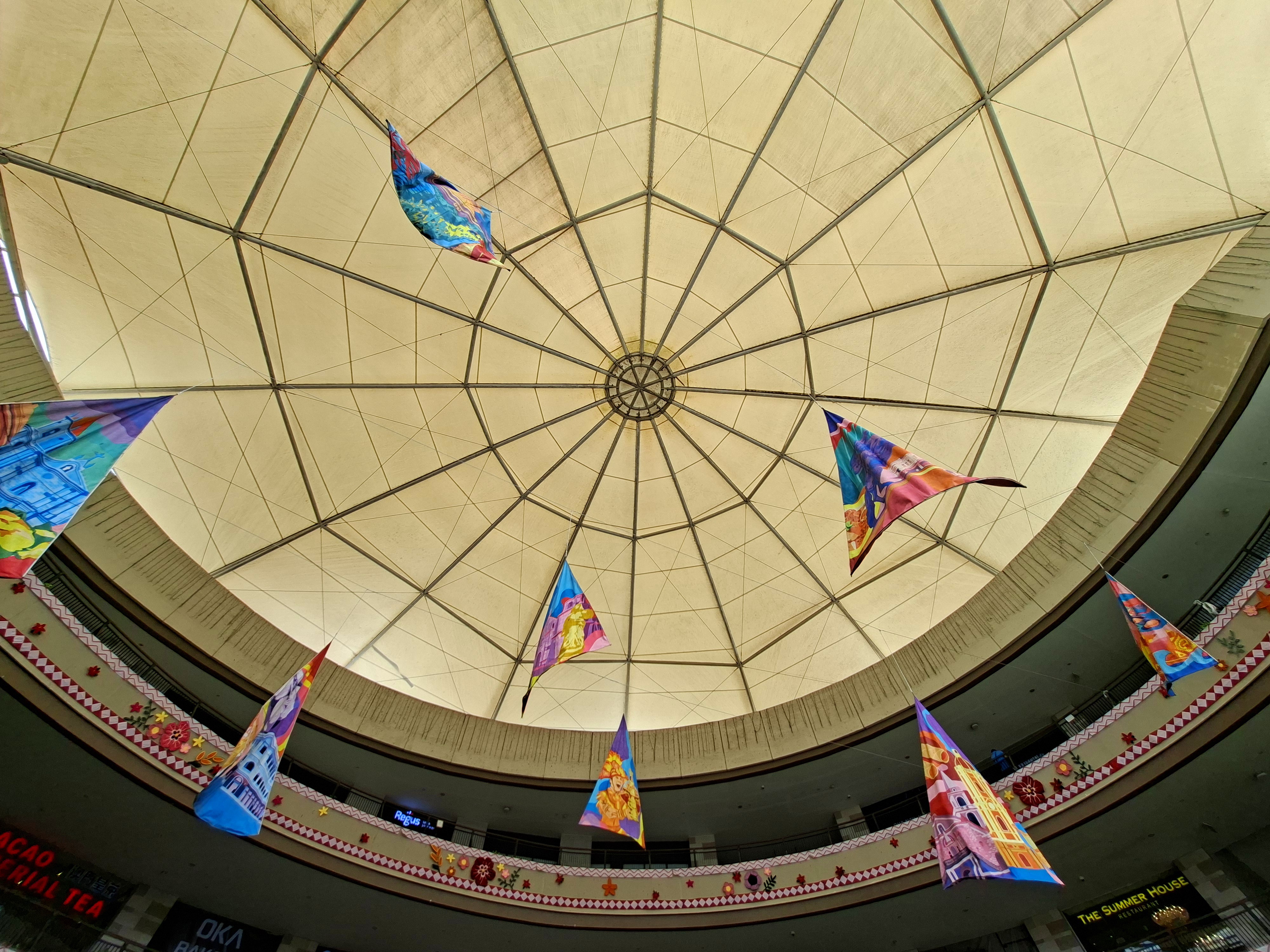 Low-angle shot looking directly up at a large fabric dome canopy ceiling with colorful triangular banners suspended beneath it inside a circular shopping mall interior.