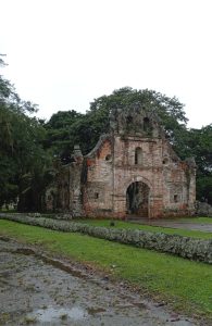 Distant view of the Ujarrás church ruins in Costa Rica, surrounded by dense trees, with a low stone wall lining the grassy path leading to the site.