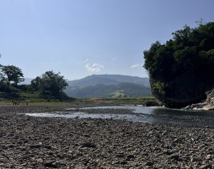 A wide, rocky riverbed with smooth grey pebbles stretching toward a shallow river. To the right, a large, tree-covered cliff overhangs the water, while lush green trees line the left bank. Mountain ranges rise under a clear blue sky in the background.
