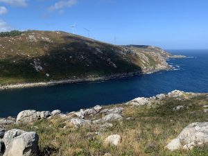 Blue sea inlet in A Barda Bay, with a green hill topped by wind turbines and rocks in the foreground.