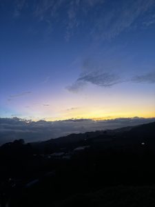 Peaceful sunset view from the mountains of Coronado, featuring a glowing yellow horizon beneath layers of clouds, a deep blue evening sky, and dark silhouettes of hills and rural buildings.
