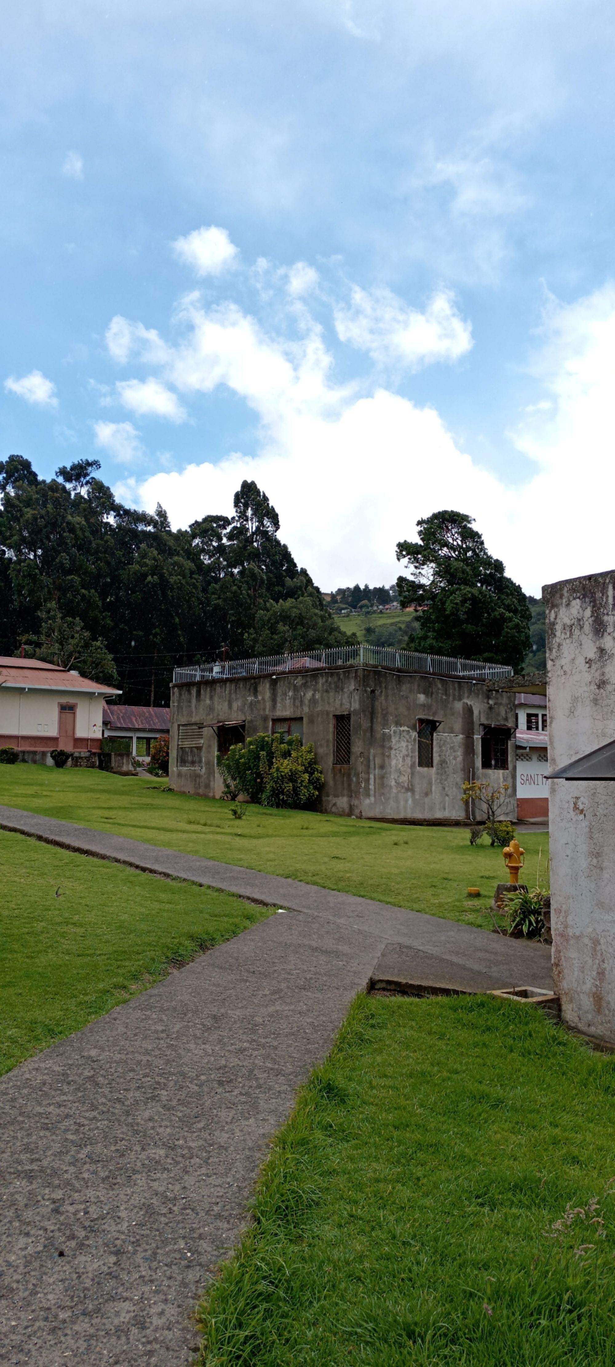 Concrete pathway leading to a weathered building with a rooftop railing at the Sanatorio Durán in Costa Rica, surrounded by open lawn and forested hills.