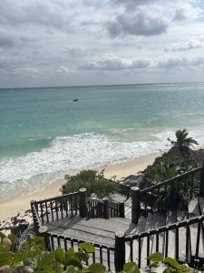 A wooden staircase winds down towards a sandy beach, surrounded by lush greenery. The ocean is a vibrant turquoise, with gentle waves lapping at the shore. In the distance, a small boat moves across the water under a cloudy sky, which features varying shades of gray.