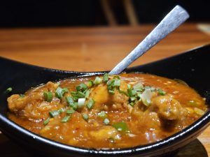 A bowl of spicy Garlic chicken gravy topped with fresh green onions. The glossy sauce, chicken pieces, and colorful garnish are captured in a close-up food composition. Location: Edavannappara, Malappuram, Kerala.
