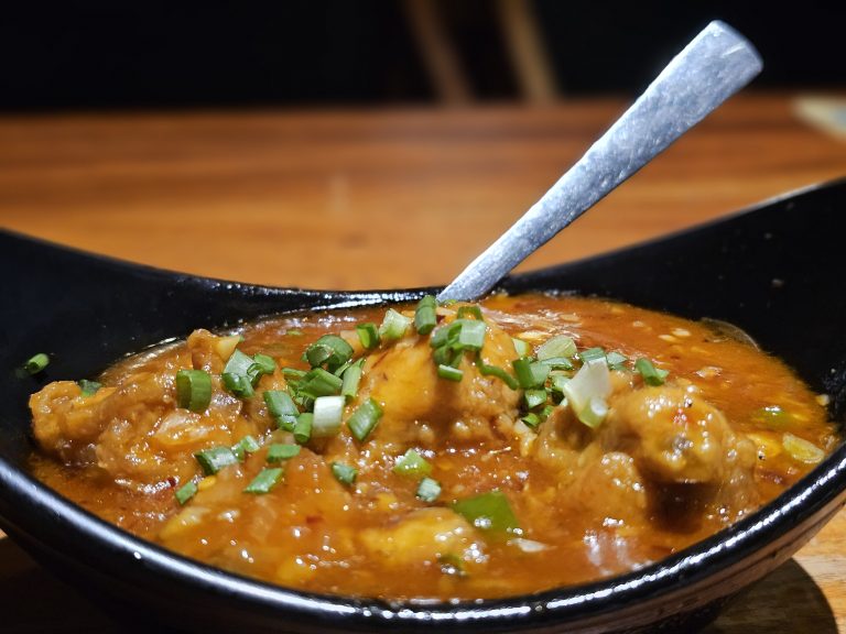 A bowl of spicy Garlic chicken gravy topped with fresh green onions. The glossy sauce, chicken pieces, and colorful garnish are captured in a close-up food composition. Location: Edavannappara, Malappuram, Kerala.