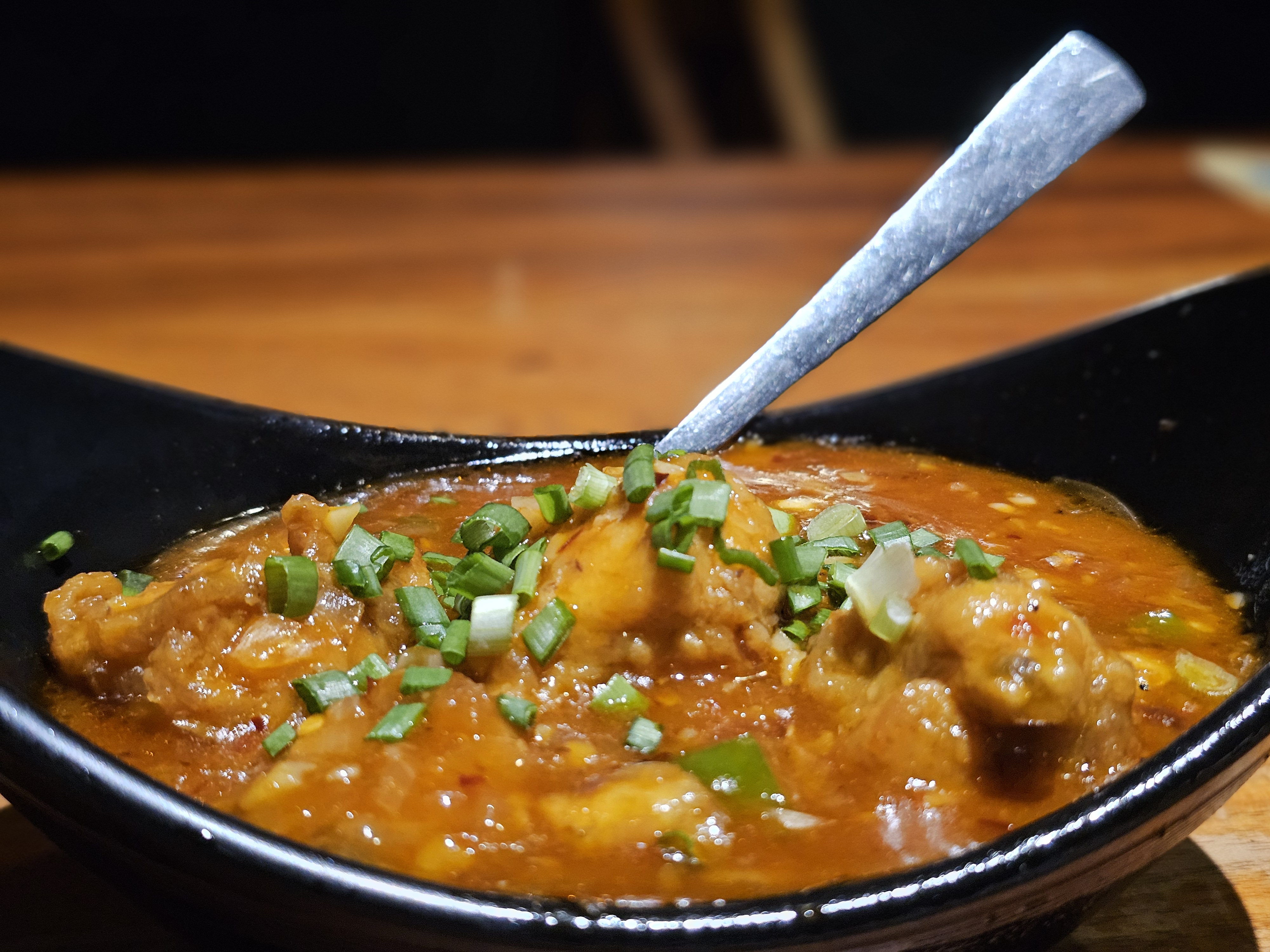 A bowl of spicy Garlic chicken gravy topped with fresh green onions. The glossy sauce, chicken pieces, and colorful garnish are captured in a close-up food composition. Location: Edavannappara, Malappuram, Kerala.