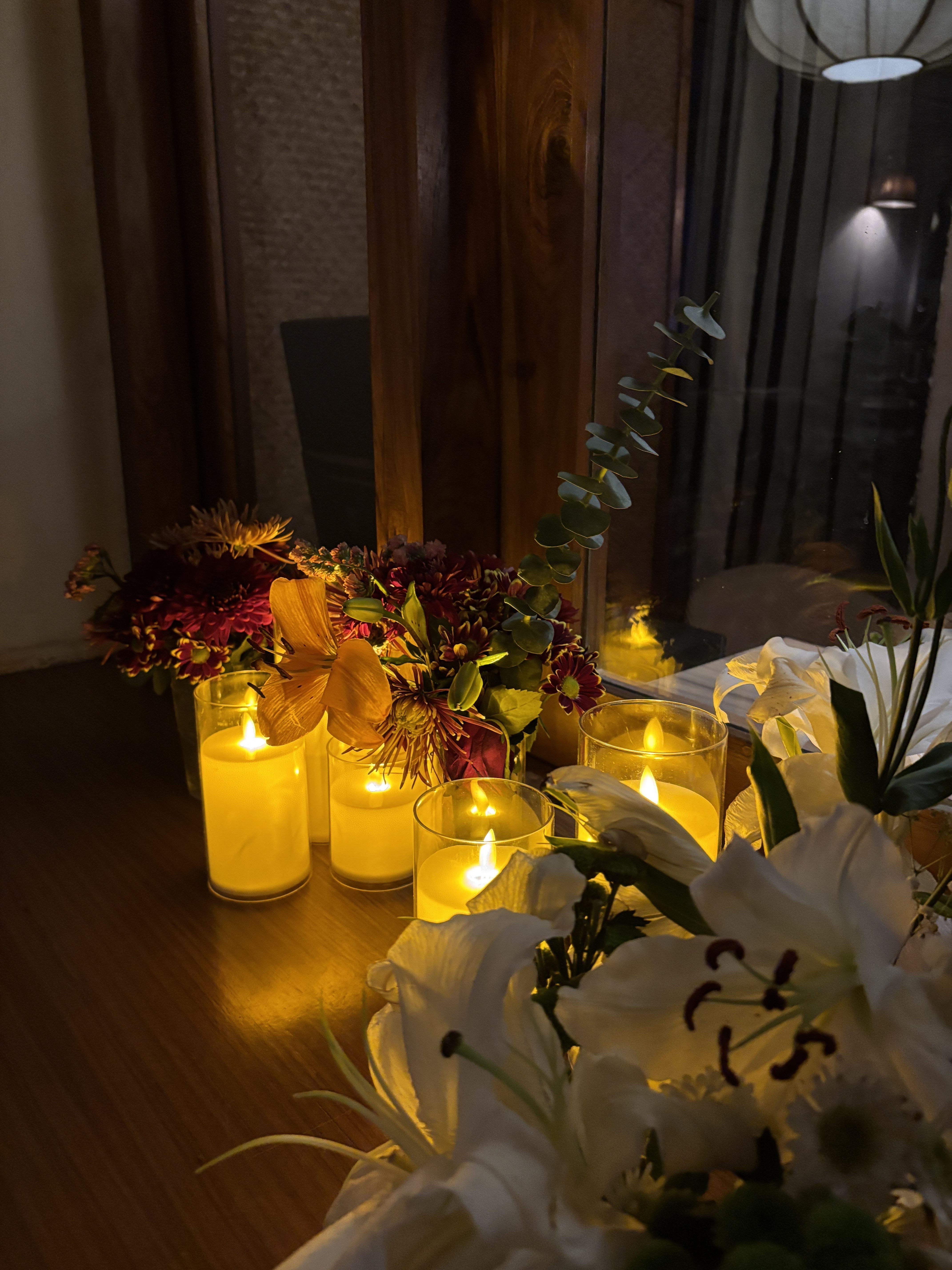 A cozy indoor arrangement of colorful flowers and white lilies sits beside a window, surrounded by glowing candles in glass holders. Warm candlelight reflects off the wooden surface, creating an intimate, peaceful atmosphere.