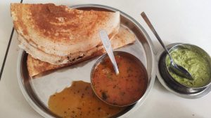 A stainless steel plate holds a stack of two golden-brown dosa, a thin and crispy Indian crepe, with visible specks of seasoning on top.
