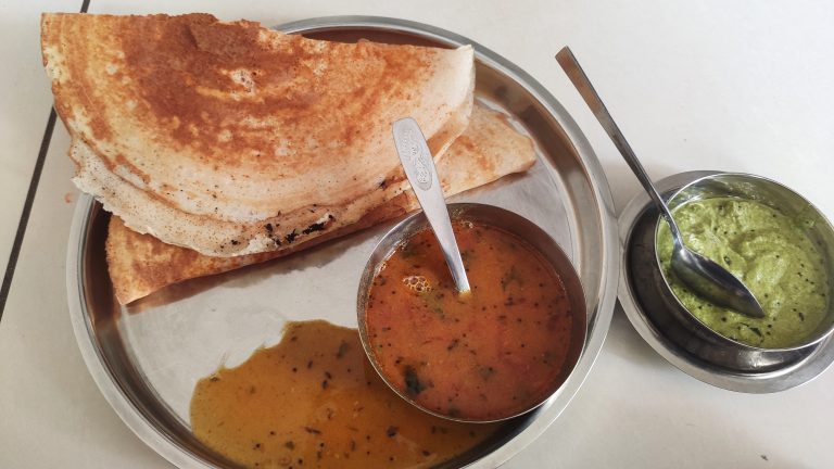 A stainless steel plate holds a stack of two golden-brown dosa, a thin and crispy Indian crepe, with visible specks of seasoning on top.
